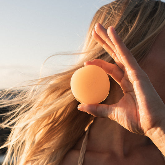 A girl with blonde surfer hair holding a bar of solid all natural conditioner after surfing.