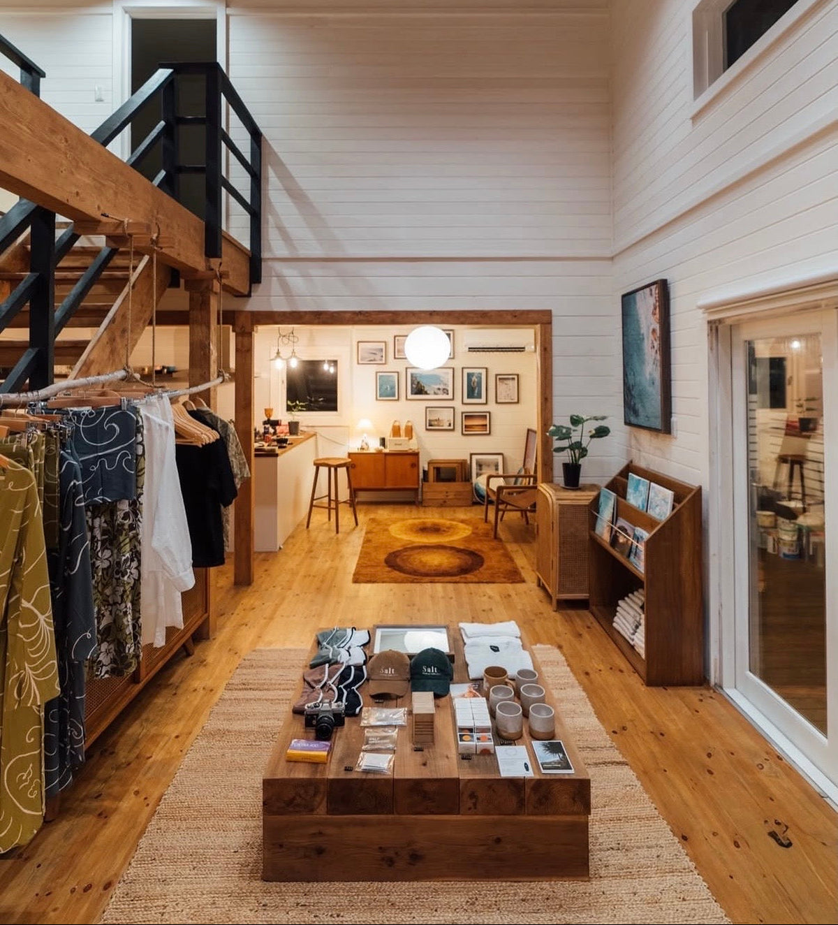 Board Butter™ surf shampoo and conditioner bars from Saltwater Cowboy displayed inside a surf shop. The space features wood-paneled walls and ceiling, a central wooden display table showcasing the all-natural surf hair care bars, a surfboard leaning against the wall, and apparel hanging on racks nearby — an example of how Board Butter™ fits into a curated surf shop retail environment, ideal for wholesale stockists looking for sustainable, plastic-free hair care.