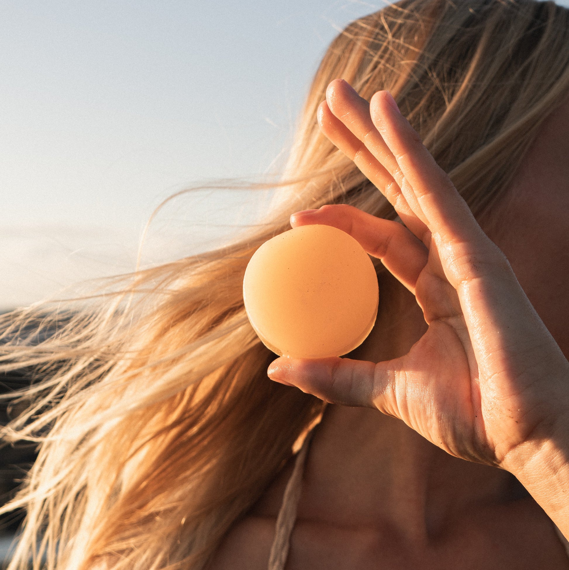 A girl with blonde surfer hair holding a bar of solid all natural conditioner after surfing.